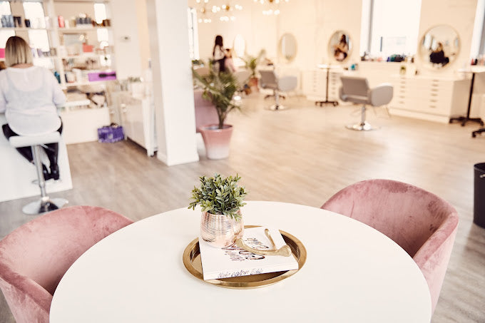 Salon view with table and pink velvet chairs in the forefront of the picture at Luxe Beauty Company.
